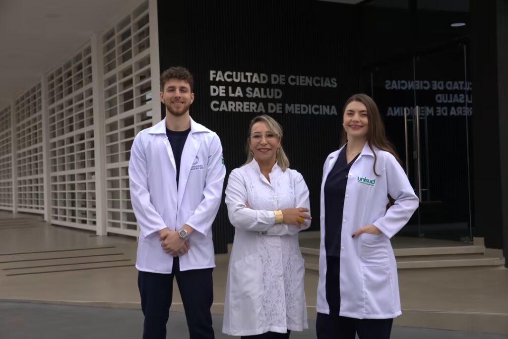 Fotografía de tres profesionales de la salud, dos mujeres y un hombre, sonriendo y de pie frente a la entrada principal del edificio de la Facultad de Ciencias de la Salud de UNINTER. Todos visten batas blancas limpias. El hombre está a la izquierda, la mujer mayor en el centro con los brazos cruzados y la mujer más joven a la derecha, quien tiene el logo de "UNINTER" visible y lleva un uniforme médico azul oscuro debajo. En la pared de fondo, de color negro con paneles, se lee en grandes letras blancas y claras: "FACULTAD DE CIENCIAS DE LA SALUD" y, justo debajo, "CARRERA DE MEDICINA". A la izquierda de la escena, se aprecian modernas celosías arquitectónicas blancas y una rampa de acceso. La imagen transmite un ambiente profesional, académico, acogedor y de pertenencia a la institución médica.