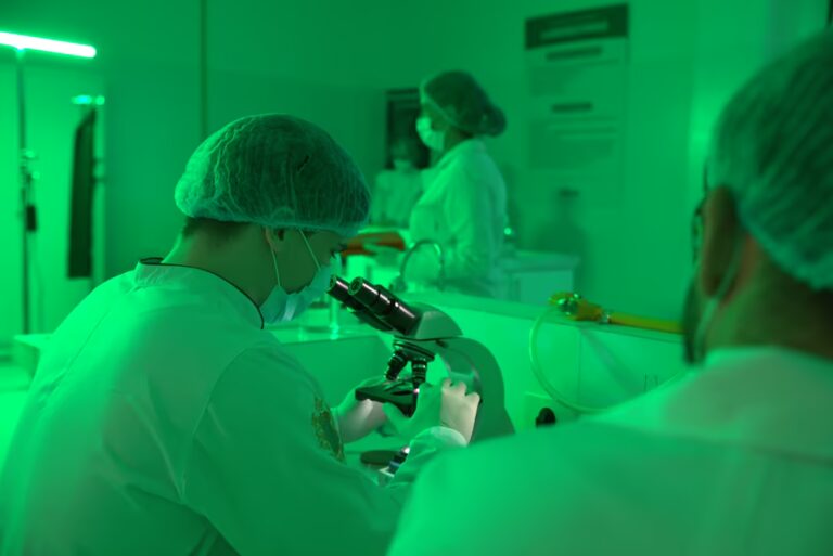 Un investigador, con bata de laboratorio, mascarilla y gorro quirúrgico, enfocado en el análisis a través de un microscopio en un laboratorio de la facultad de medicina. Se ve a otros colegas trabajando en el fondo, todo bajo una luz ambiental verde constante.