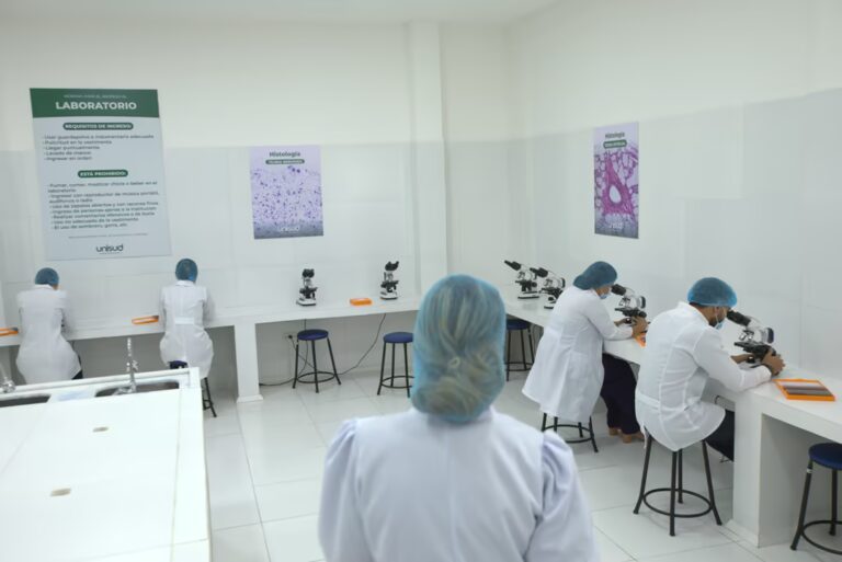 Vista trasera de una instructora y varios estudiantes de medicina de UNISUD, todos vestidos con batas blancas y cofias azules, trabajando con microscopios en un laboratorio de histología luminoso y limpio. Las paredes tienen carteles con normas del laboratorio y muestras de tejido, y el logo de UNISUD es visible en sus batas y materiales.