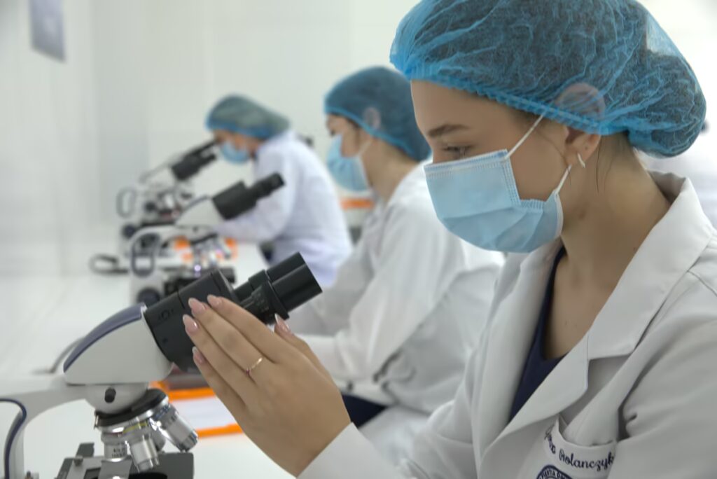 Fotografía de una clase práctica en un laboratorio de anatomía médica de una institución de enseñanza superior. Un instructor con bata de laboratorio, mascarilla y gorro quirúrgico explica una lección de anatomía junto a un detallado modelo anatómico de cuerpo completo (esqueleto y músculos) y una pizarra con anotaciones. Estudiantes en primer plano y sentados alrededor de mesas de acero inoxidable, también con batas, mascarillas y gorros quirúrgicos, observan atentamente la presentación. Al fondo se aprecian vitrinas con otros modelos médicos y equipos de laboratorio. El entorno es limpio, profesional y académico.