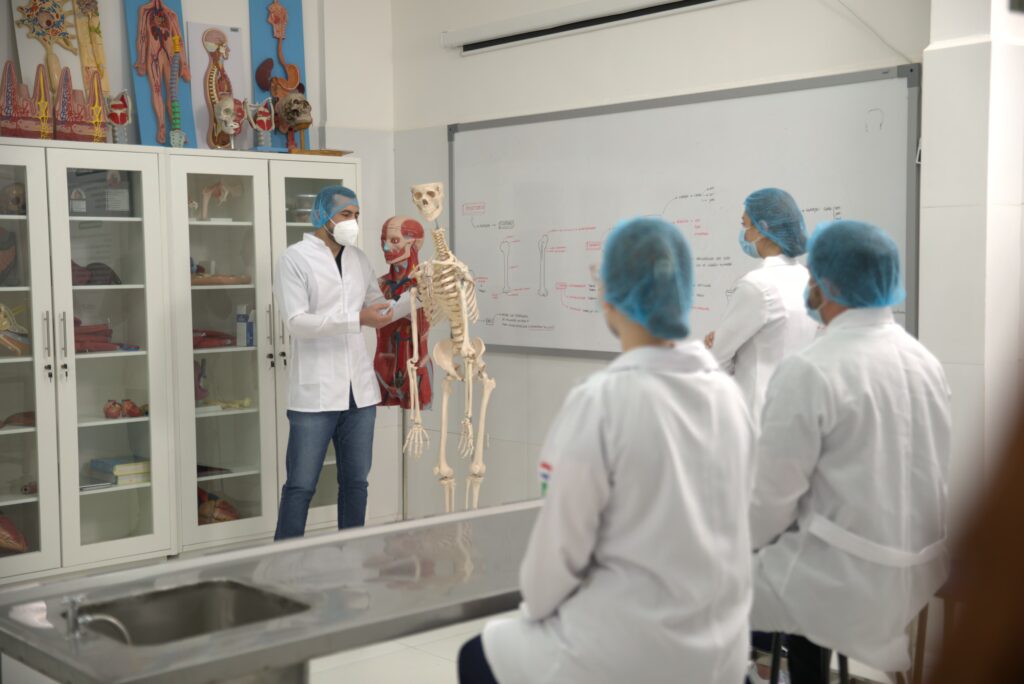 Un instructor de medicina, con bata de laboratorio, mascarilla N95 y gorro quirúrgico, señala un modelo de esqueleto humano mientras explica algo a un estudiante desenfocado en primer plano. Están en un aula de anatomía con vitrinas de vidrio llenas de diversos modelos anatómicos (corazones, cráneos, columna vertebral) y una pizarra con diagramas de anatomía en español.