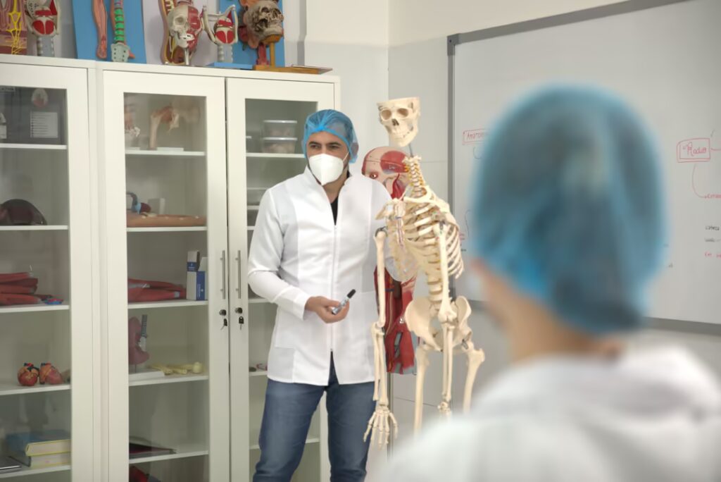 Un instructor de medicina, con bata de laboratorio, mascarilla N95 y gorro quirúrgico, señala un modelo de esqueleto humano mientras explica algo a un estudiante desenfocado en primer plano. Están en un aula de anatomía con vitrinas de vidrio llenas de diversos modelos anatómicos (corazones, cráneos, columna vertebral) y una pizarra con diagramas de anatomía en español.