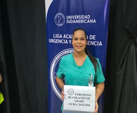 Estudiante de UNISUD sosteniendo un cartel de la colación de grado frente al banner de la Liga de Urgencia y Emergencia.