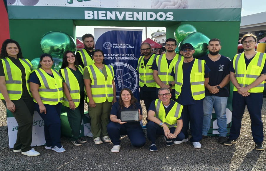 Equipo de estudiantes voluntarios con chalecos amarillos posando con una placa durante el evento Med Start Unisud.