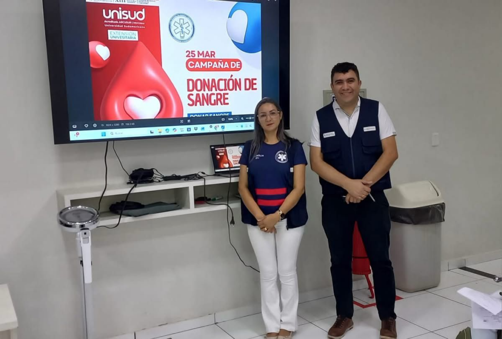 Docentes encargados frente a la pantalla promocional durante la jornada de donación de sangre en UNISUD.