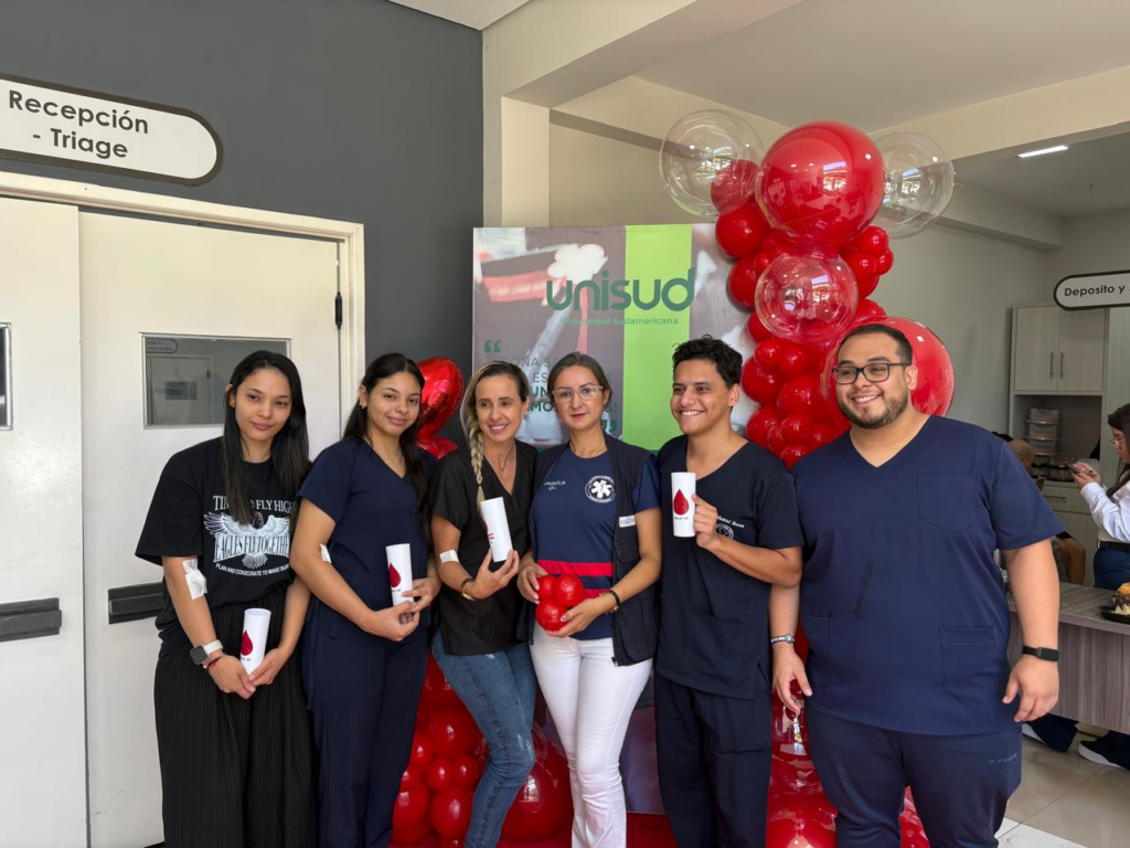 Organizadores en la jornada de donación de sangre de UNISUD posando junto a un cartel temático con globos rojos.
