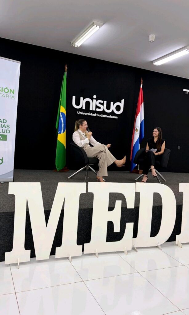 Dos mujeres conversando en el escenario durante la jornada de medicina UNISUD frente a banderas de Brasil y Paraguay.