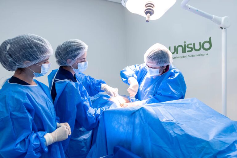 Fotografía detallada que captura una escena de instrucción clínica o cirugía práctica en un quirófano estéril de la Universidad Sudamericana (unisud). Tres profesionales de la salud, completamente vestidos con batas quirúrgicas azules, mascarillas, gorros y guantes, están presentes. Dos de ellos en el centro de la imagen están inclinados sobre un paciente cubierto por sábanas quirúrgicas azules, realizando activamente un procedimiento quirúrgico bajo una potente lámpara de quirófano. El tercer profesional, también con vestimenta quirúrgica completa, observa atentamente en primer plano a la izquierda. El logotipo verde y el texto 'unisud' y 'Universidad Sudamericana' son visibles en la pared del fondo a la derecha, confirmando la ubicación institucional.